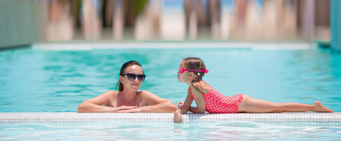Frau mit Sonnenbrille im Freibad, Mädchen liegt am Beckenrand mit pinkem Badeanzug und Schwimmbrille
