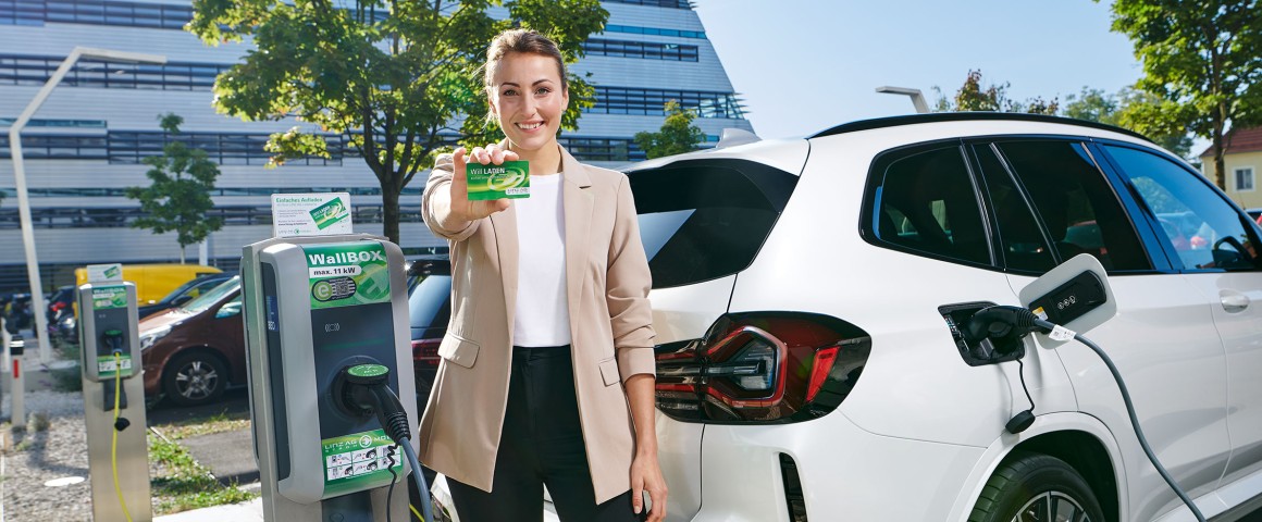 Eine Dame im Business-Look steht vor einem modernen Bürogebäude und zeigt am Parkplatz neben einer E-Ladesäule eine Ladekarte.