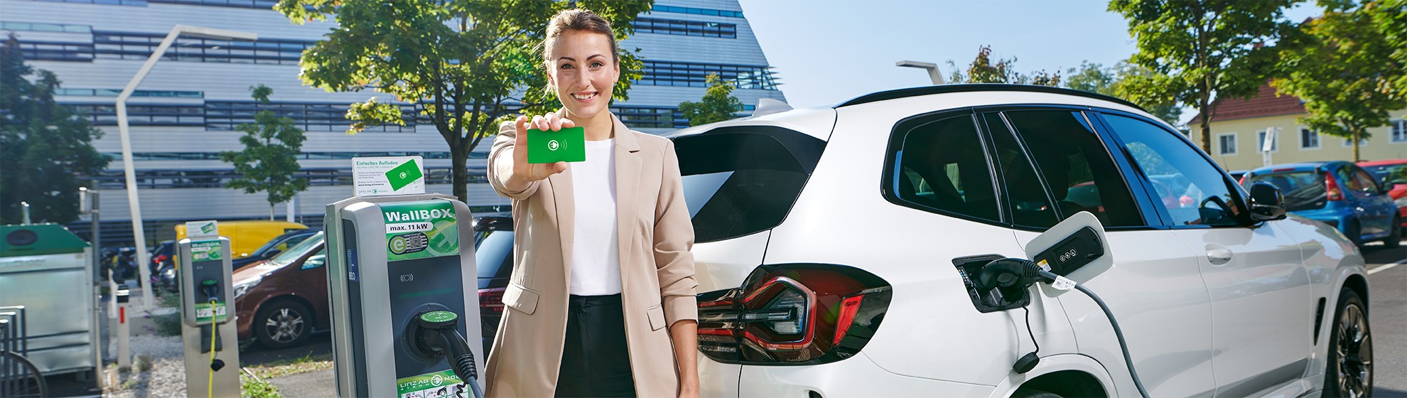 Eine Dame im Business-Look steht vor einem modernen Bürogebäude und zeigt am Parkplatz neben einer E-Ladesäule eine Ladekarte.