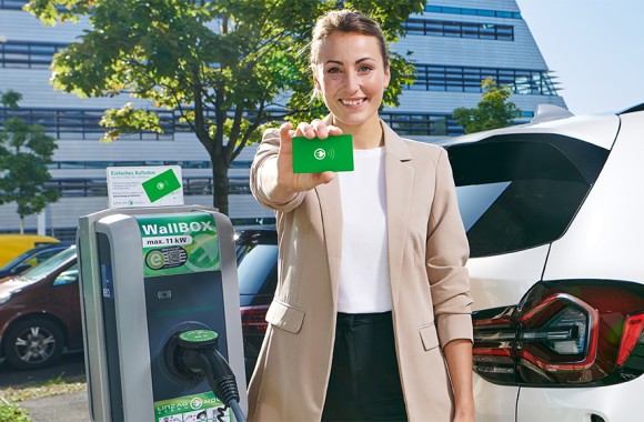 Eine Dame im Business-Look steht vor einem modernen Bürogebäude und zeigt am Parkplatz neben einer E-Ladesäule eine Ladekarte.