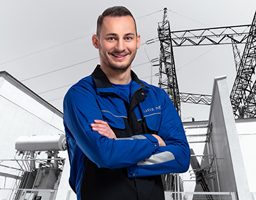 Ein Facharbeiter der Linz AG in der Arbeitskleidung lächelt in die Kamera, im Hintergrund ist ein Umspannwerk zu sehen.