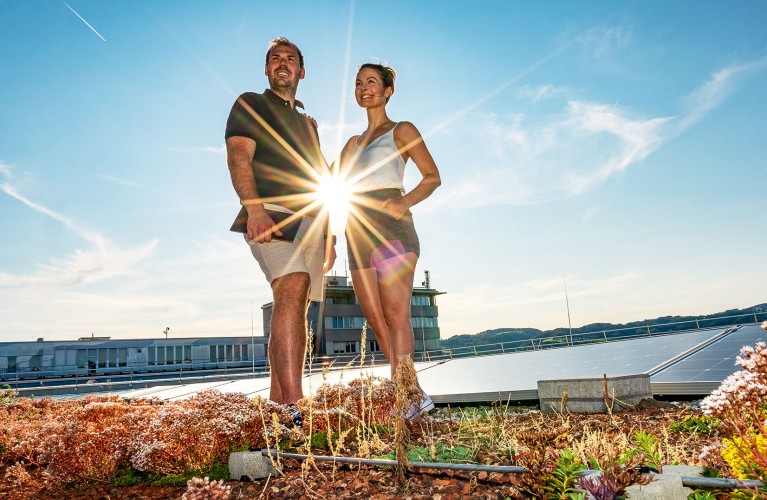 Ein Mann mit einem Tablet in der Hand und eine Frau stehen nebeneinander vor einer Photovoltaikanlage und die Sonne scheint hinter ihnen.