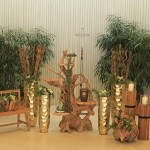 Urne mit Baumsymbol steht auf dekorativem Baumwurzel-Gebilde. Links und rechts daneben stehen jeweils zwei große goldene Vasenmit Pflanzen daneben. Weiters sieht man rechts daneben eine Holzbank mit Blumenschmuck. Im Hintergrund sind Pflanzen. 