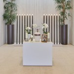 Die Urne steht auf weißem Podest mitten im Raum, daneben ein Foto des*der Verstorbenen und weiße Orchideen.