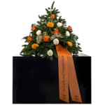 grünes Bukett mit unterschiedlichen orangenen Blumen und weißen Nelken und orangener Schleife mit Aufschrift "in Liebe und Dankbarkeit" steht auf schwarzem Podest.