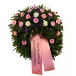 grüner Kranz mit rosa Gerbera, lila Nelken und weißen Rosen, dazu eine rosa Schleife mit Aufschrift "Unsere Liebe wird Dich für immer begleiten"