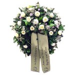 grüner Kranz mit unterschiedlichen weißen Blumen und lila Akzenten. Dazu eine beige Schleife mit Aufschrift "in lieber Erinnerung Anna und Walter, Regina und Klaus"