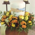 Blumenkranz mit unterschiedlichen gelben und orangenen Blumen rund um die Urne