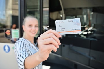 Frau steht lächelnd vor Straßenbahn und hält MEGA-Ticket in die Kamera.