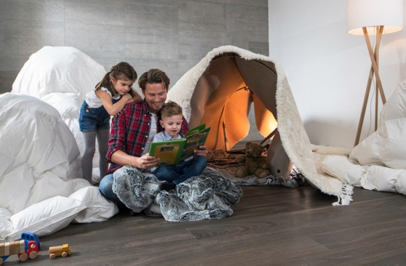 Ein Vater sitzt mit seinen beiden Kindern im Wohnzimmer am Boden und blättert einen Bildband durch. Im Hintergrund ein selbstgebautes Zelt aus Bettlaken und Decken.